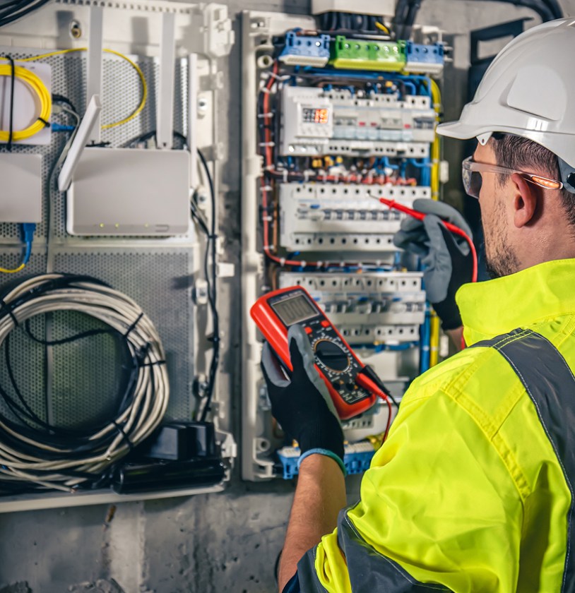 A technician in a hard hat and reflective shirt uses a multimeter on an electrical panel with various wires and devices.