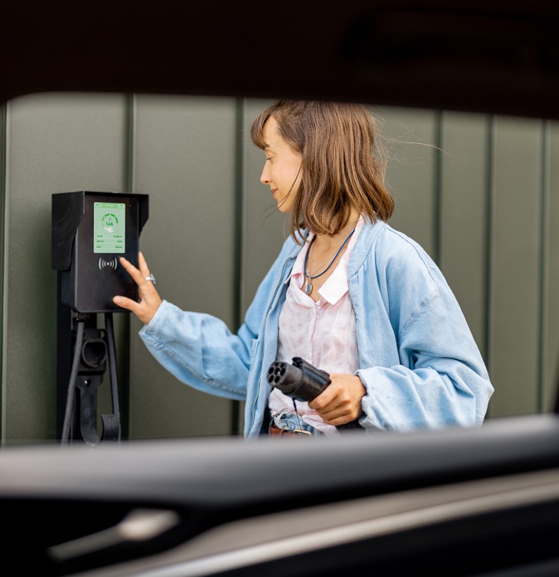 A person interacts with an electric vehicle charging station, holding a charging plug, with a green wall in the background.